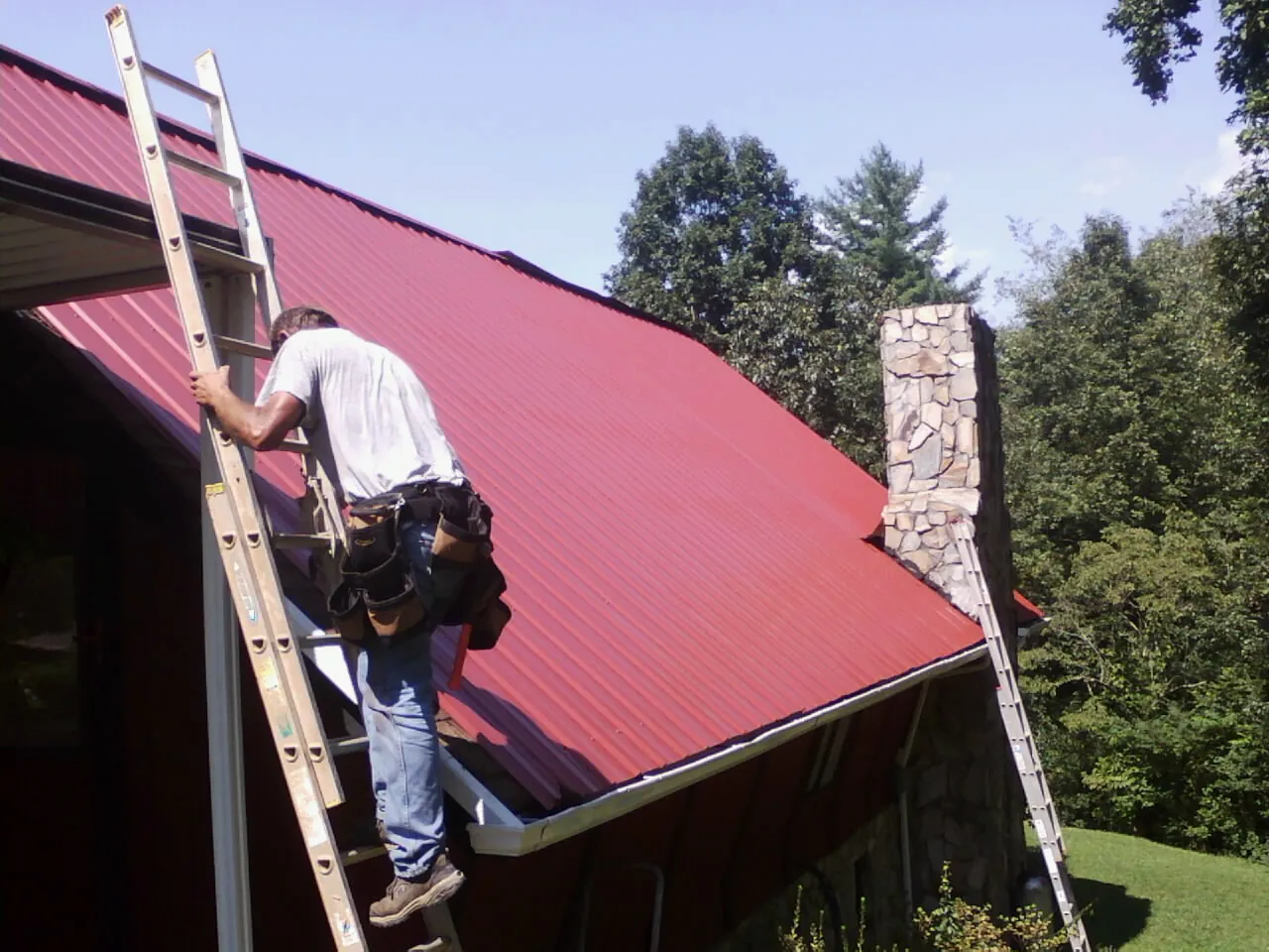 Roofer on a ladder beside a red metal roof and stone chimney — Soffit & Fascia Repair by Roofing Brothers in Brantingham